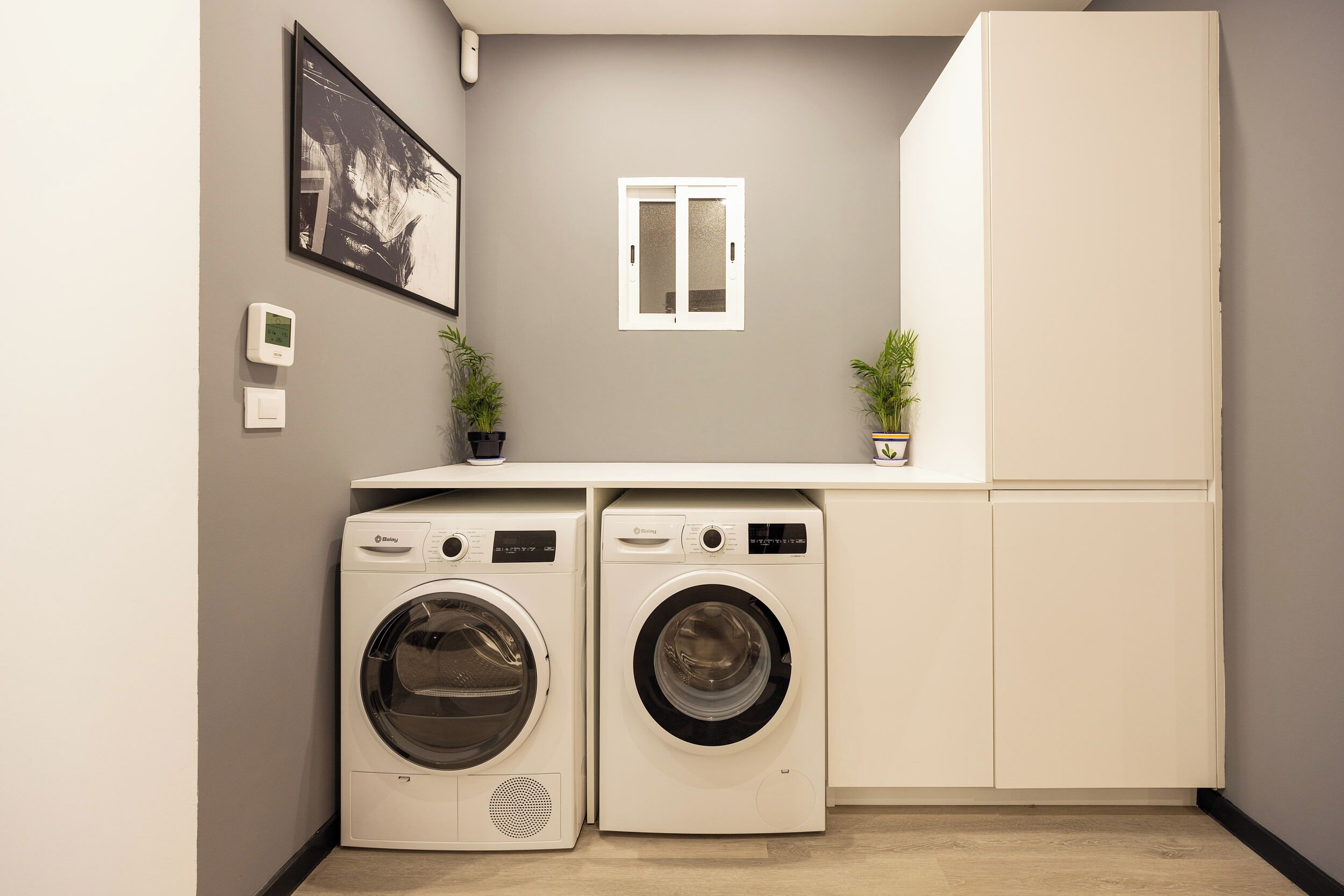 Un espace buanderie compact comprenant deux machines à laver à chargement frontal, une petite fenêtre et une plante en pot.
