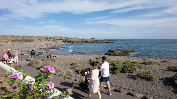 Una playa con gente, un cochecito de bebé y una costa rocosa.