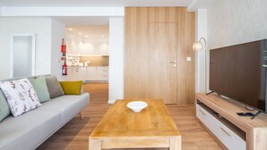 A modern living room with a wooden coffee table, a sofa with cushions, a flat-screen TV on a wooden stand, and a kitchen in the background.