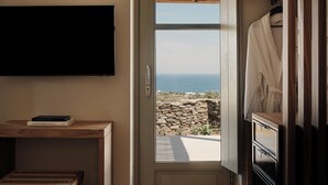 A room with a television mounted on the wall, a wooden table with books, and a door leading to a balcony with a view of the sea. - Stamna Sifnos, a member of Design Hotels (Sifnos)