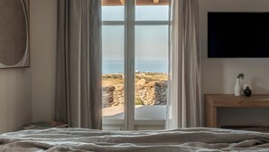 A bedroom with a large window, a bed, a bedside table with a vase, and a mounted TV. - Stamna Sifnos, a member of Design Hotels (Sifnos)