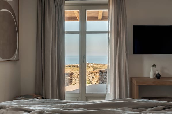 A bedroom with a large window, a bed, a bedside table with a vase, and a mounted TV. - Stamna Sifnos, a member of Design Hotels (Sifnos)