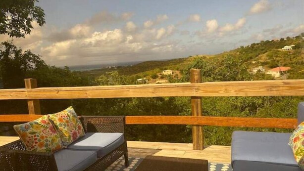 A wooden deck with wicker furniture and cushions, overlooking a hilly landscape with houses. - The Cottage at Lowry Hill (Christiansted)