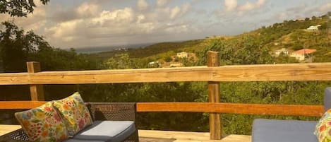 Una terraza de madera con mobiliario de mimbre y cojines, con vistas a un paisaje montañoso con casas.