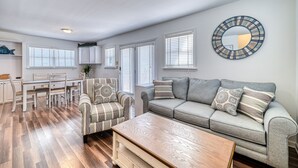 A modern living room with a grey sofa, striped armchair, and a wooden coffee table. There is a dining area with a white table and chairs, and a kitchen area with cabinets and a countertop. - Ocean-View Retreat, Sleep 8, 6-Minute Walk to Restaurants, Pet friendly (Surf City)