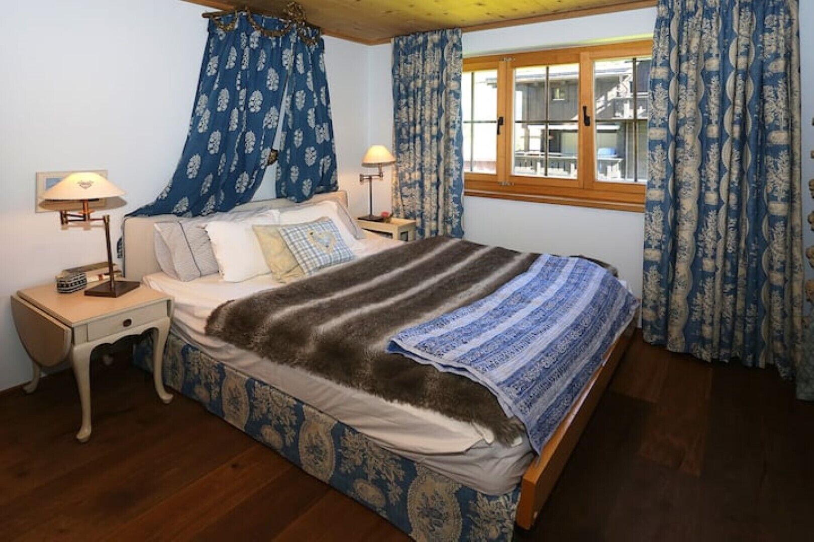 A bedroom with a bed, two bedside tables, a window with curtains, and a wooden ceiling.