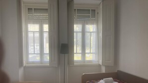 Habitación con dos camas, una con cubrecama blanco y la otra con cubrecama estampado y de colores. Entre las camas hay una lámpara de pie, y a la izquierda, una ventana con persianas.