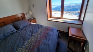 A bedroom with a bed, a nightstand, a lamp, and a window with a mountain view. - Habitamar El Colorado (Lo Barnechea)