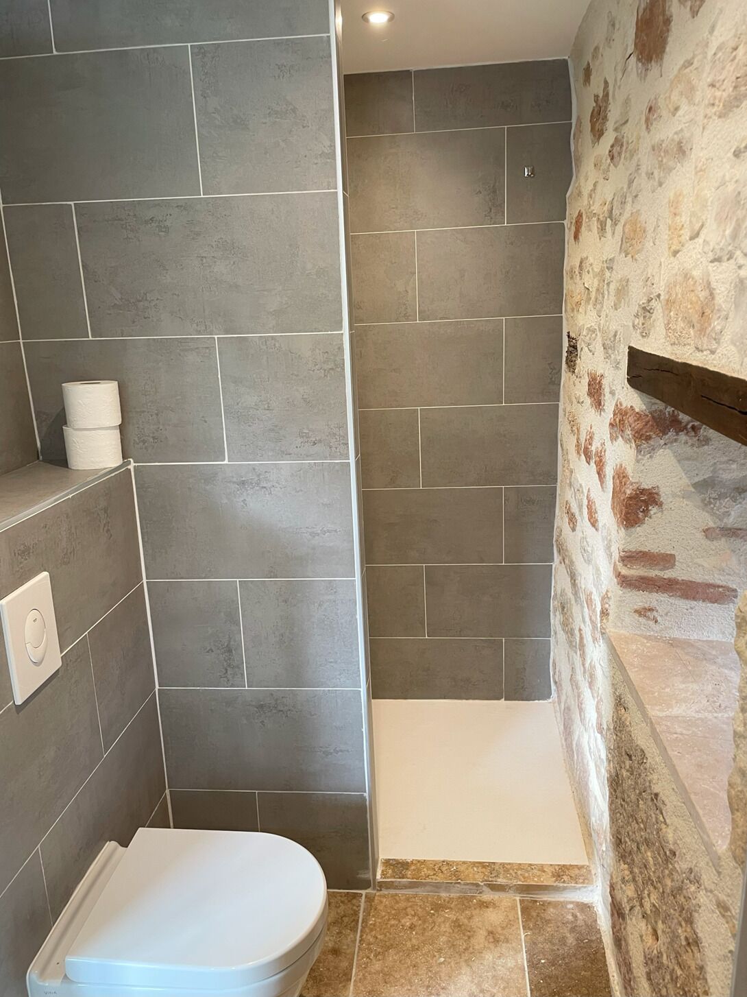 Casa de banho com azulejos cinzentos, sanita branca e zona de duche com paredes de pedra.