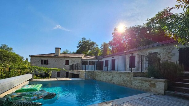 A swimming pool with a wooden deck, stone buildings, and a clear sky.