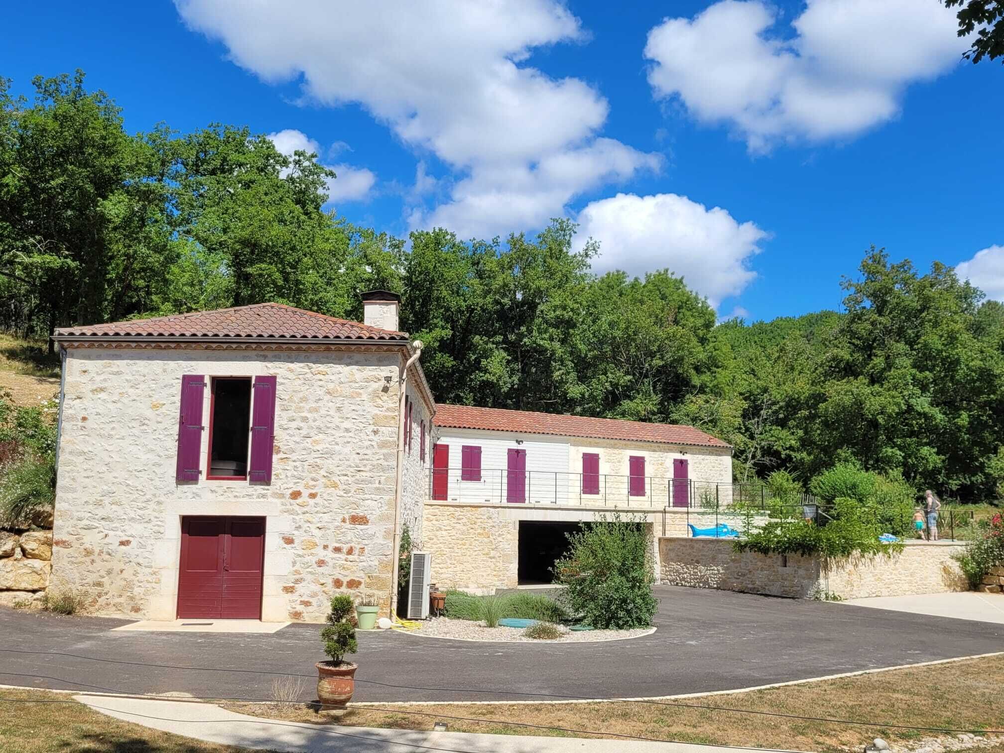 A stone building with a red door and shutters, a covered driveway, and a small garden area.