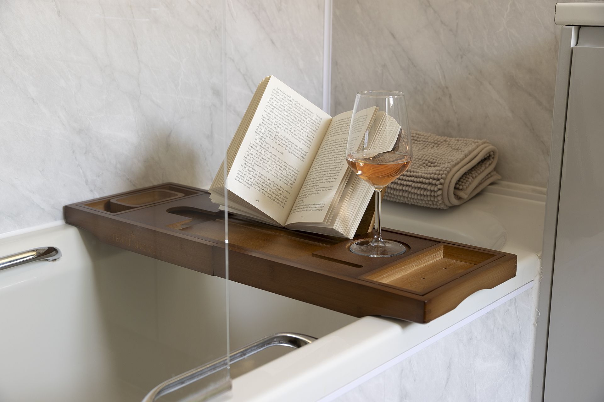 A bathroom with a wooden tray holding an open book and a glass of wine.