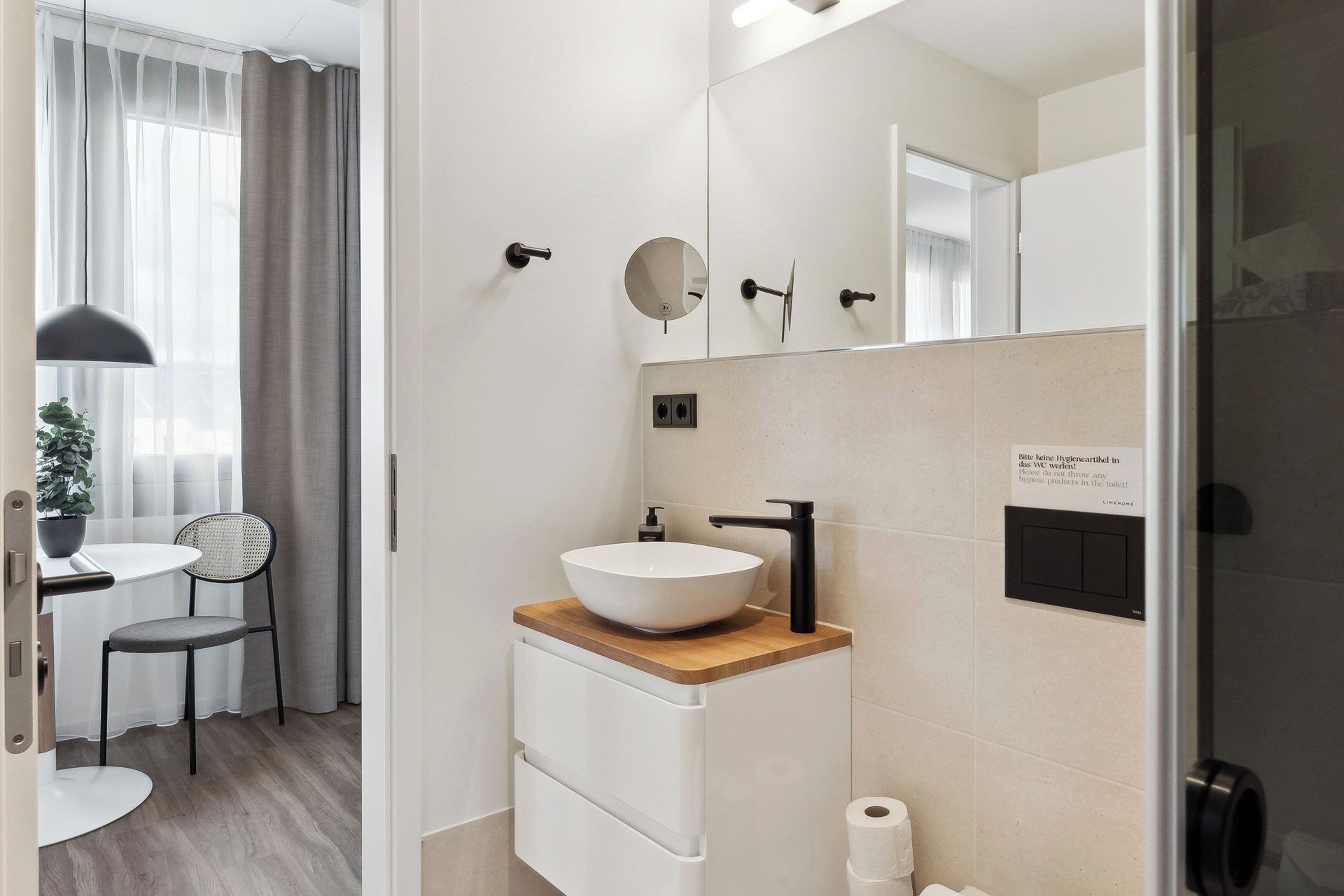 Une salle de bain moderne avec un lavabo blanc, un plan de toilette en bois et un miroir au-dessus. Une petite table et une chaise se trouvent dans la pièce adjacente.