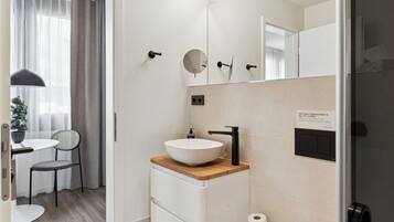 A modern bathroom with a white sink, wooden countertop, and a mirror above it. There is a small table and chair in the adjacent room.