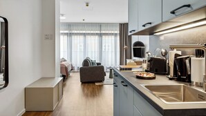 Una cuina moderna amb forn integrat, màquina de cafè i un rentaplat. Hi ha una zona de menjador amb taula i cadires, i un sofà al fons.