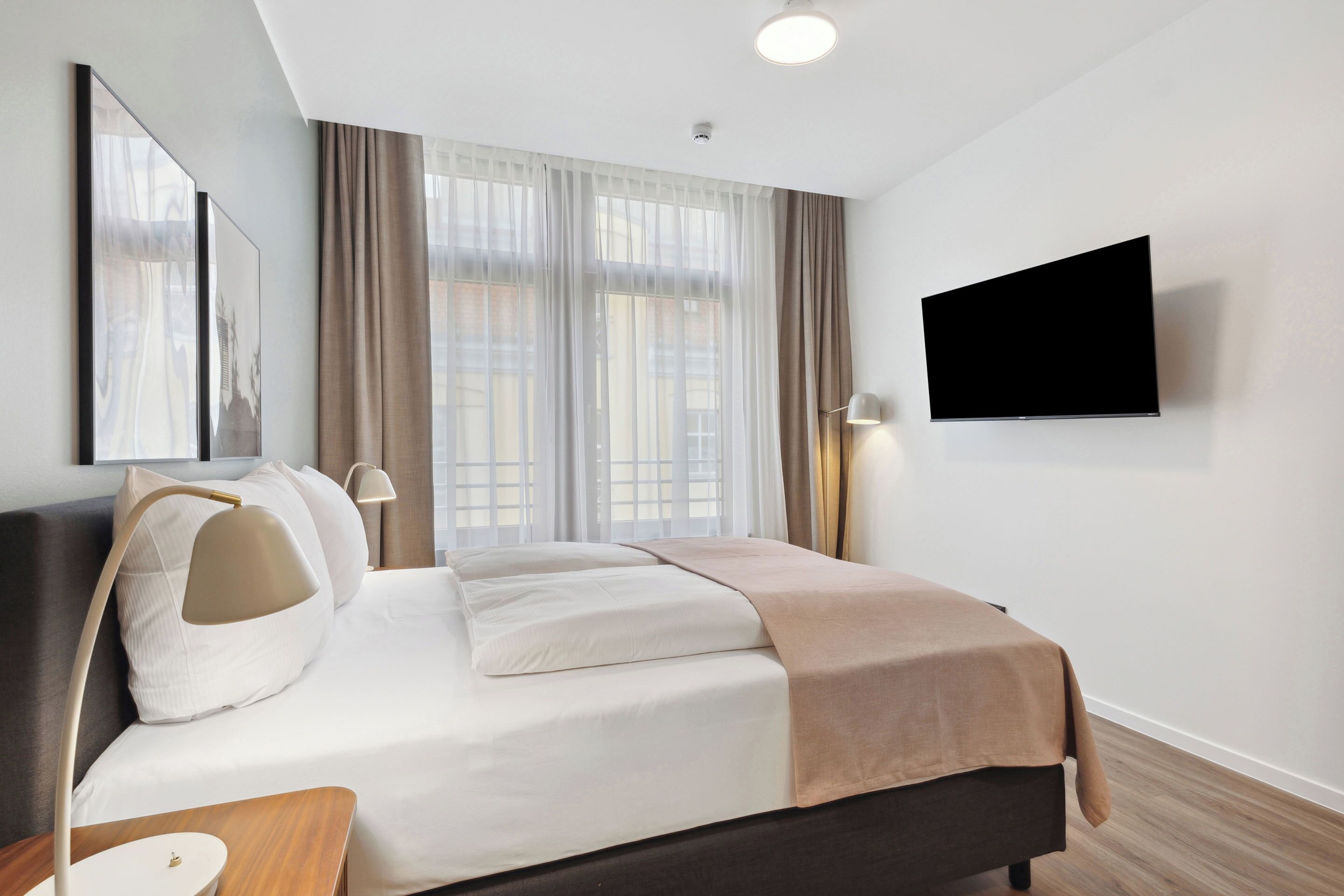 Une chambre d’hôtel moderne dotée d’un grand lit, d’un téléviseur à écran plat fixé au mur et d’une grande fenêtre avec des rideaux légers.