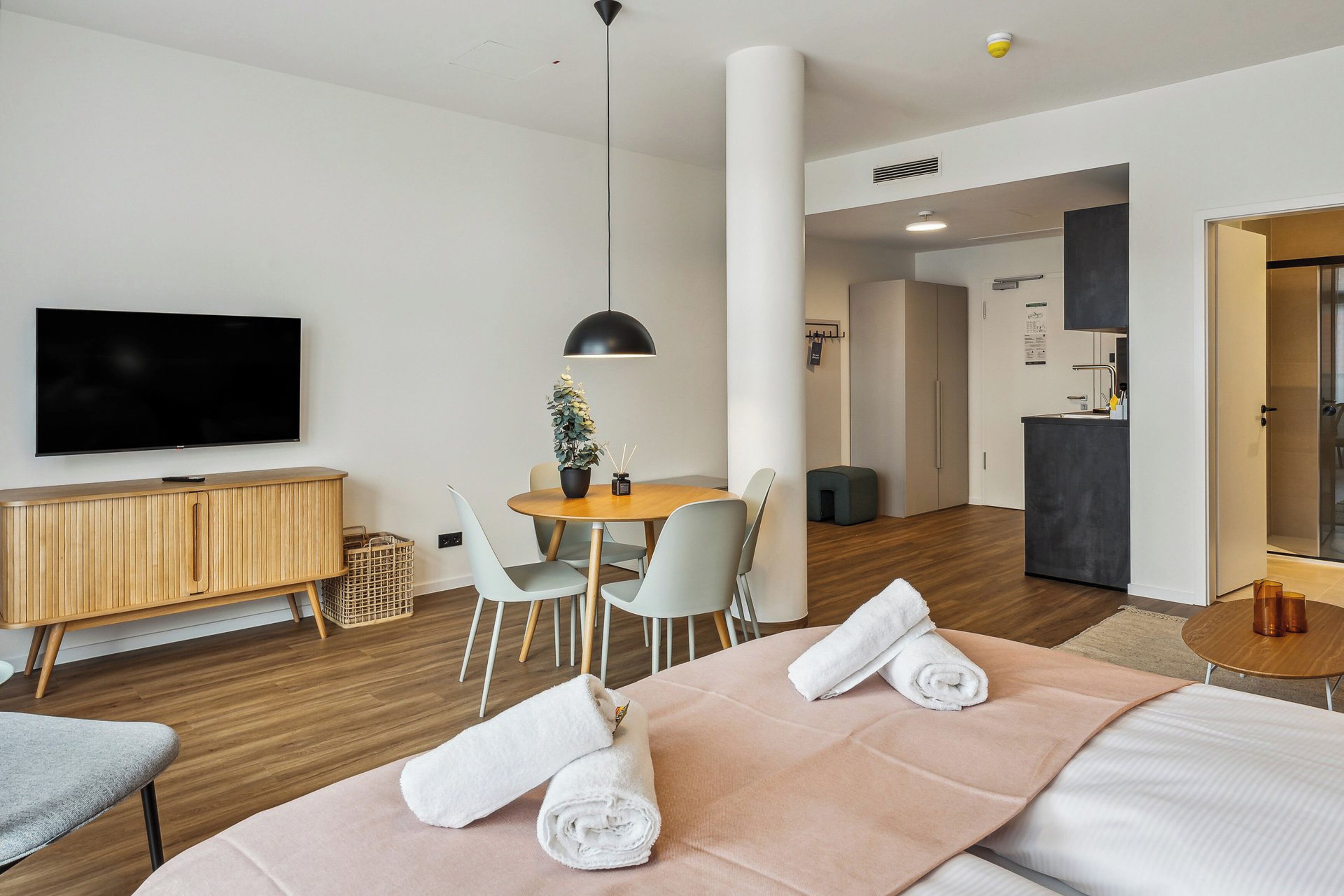 A modern hotel room with a bed, a TV, a dining table, and a small kitchen area.