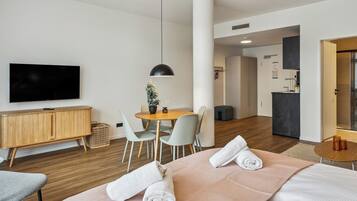 A modern hotel room with a bed, a TV, a dining table, and a small kitchen area.