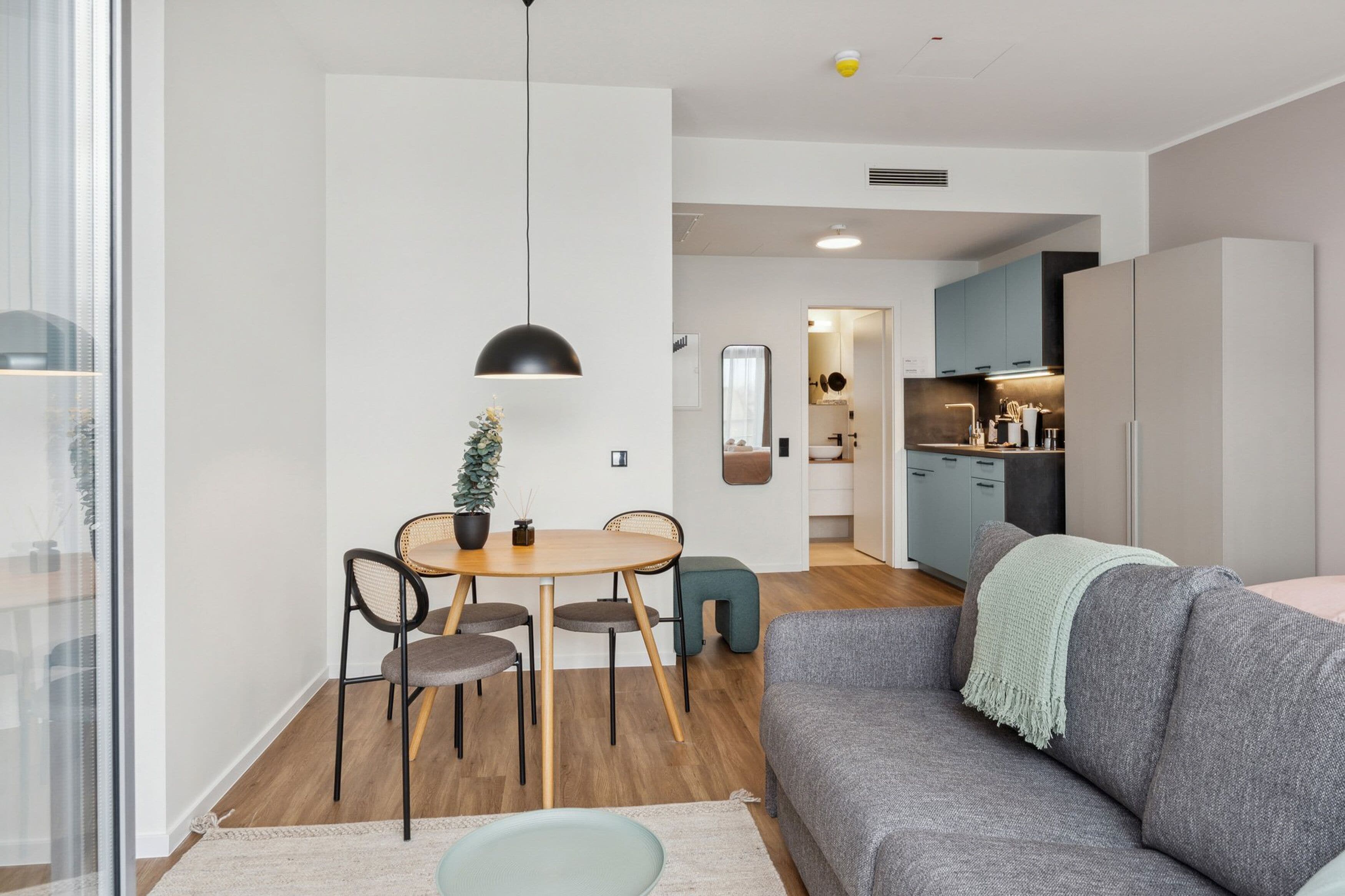 Una sala de estar moderna con un sofá gris, una mesa redonda de comedor con cuatro sillas, una pequeña planta en maceta y una lámpara colgante.