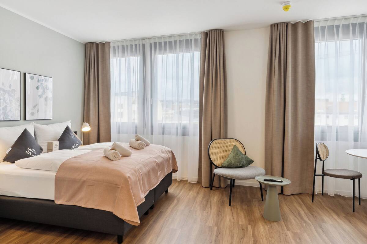 A modern hotel room with a large bed, two chairs, a small table, and a view of the city through large windows.