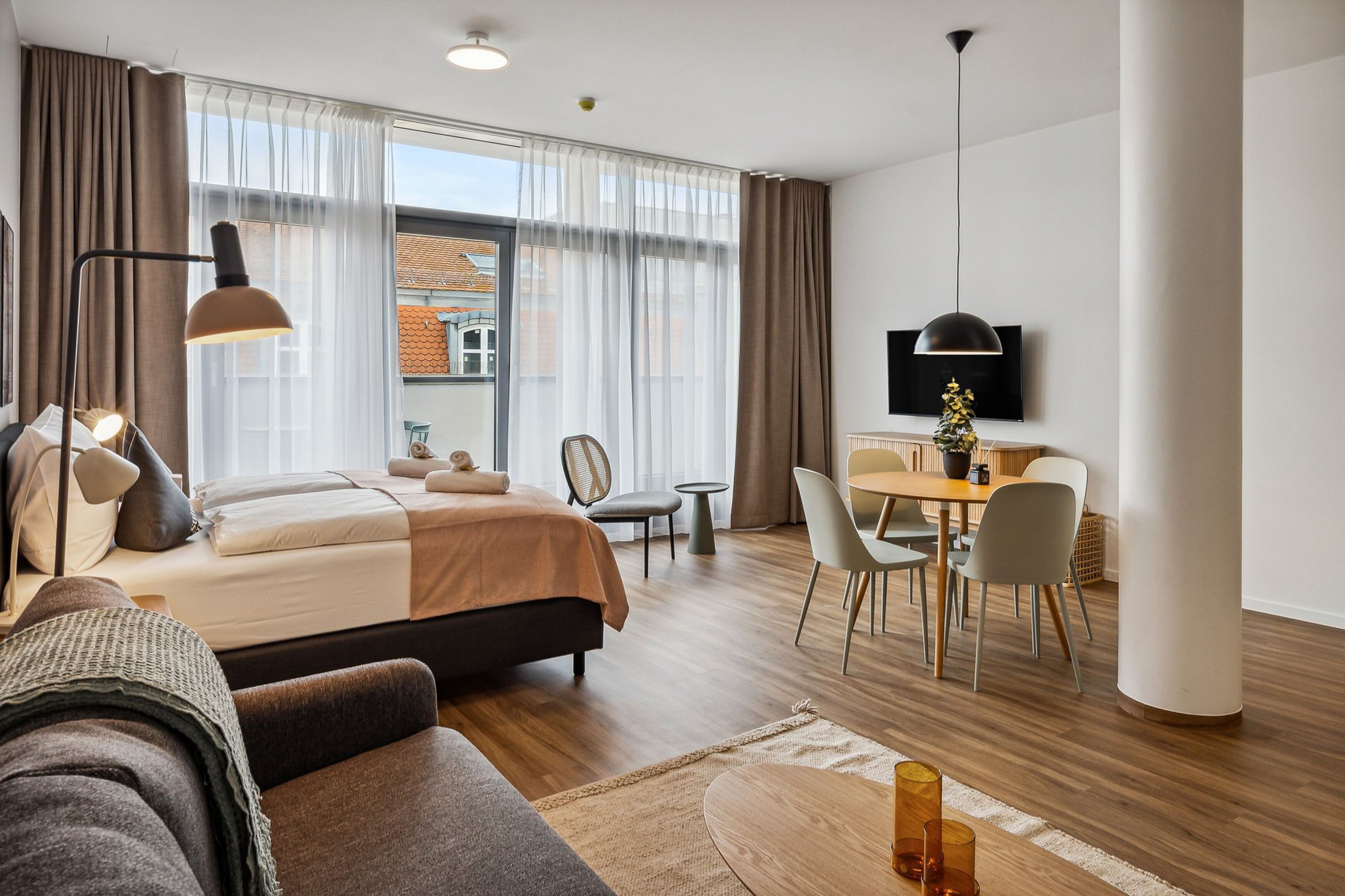 A modern hotel room with a bed, a sofa, a dining area, and a TV.