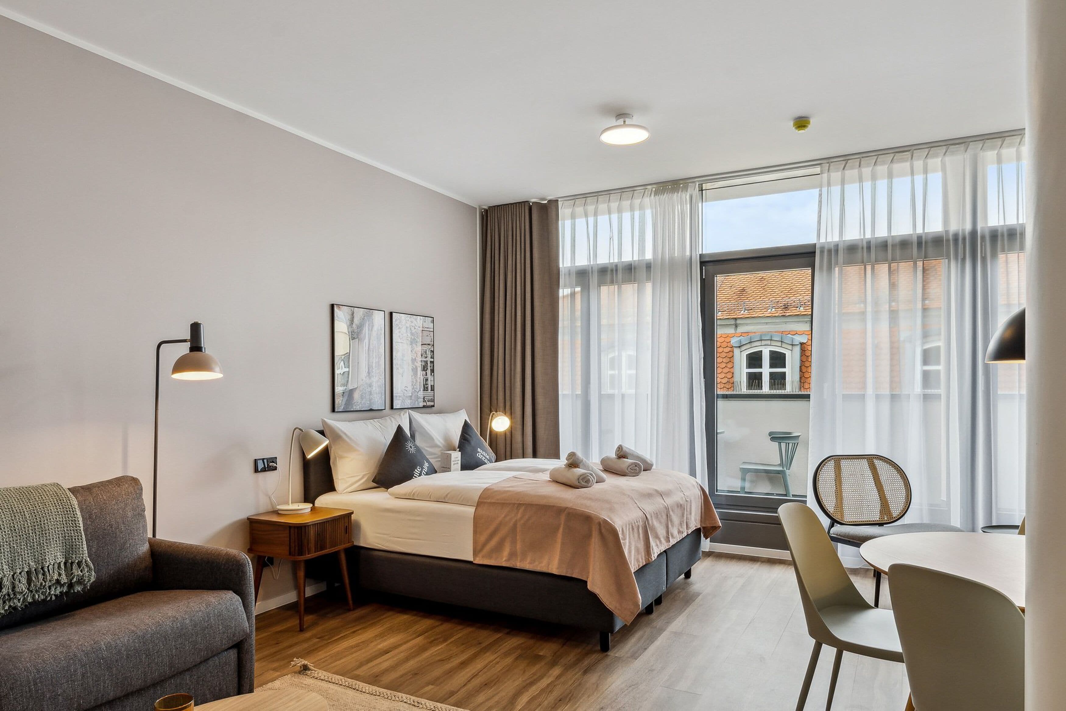 Una camera da letto moderna con un letto, comodini, una scrivania e una sedia. Sono presenti tende, una finestra e vista sugli edifici.