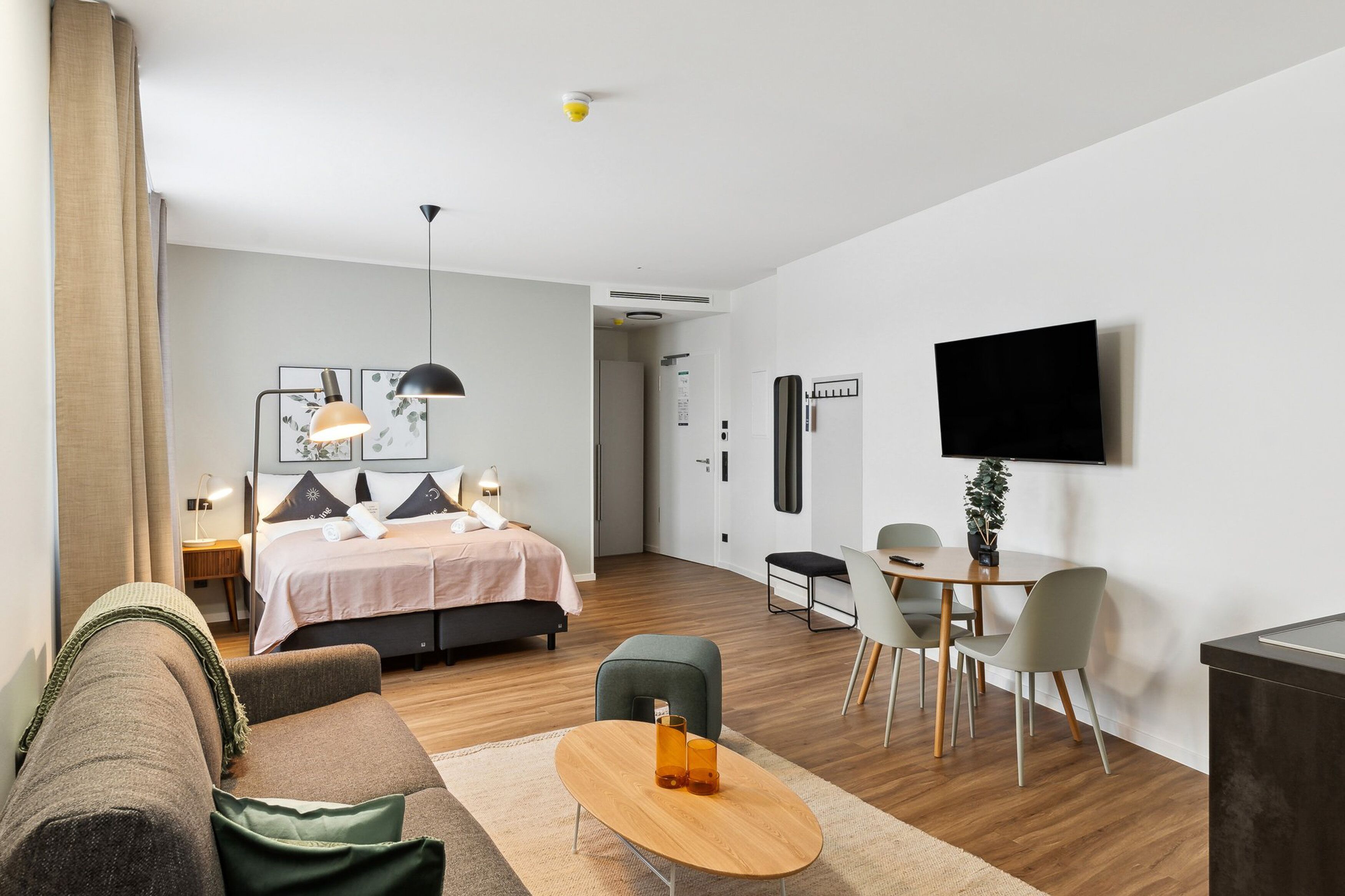 A modern hotel room with a bed, a desk, a dining area, and a TV mounted on the wall.