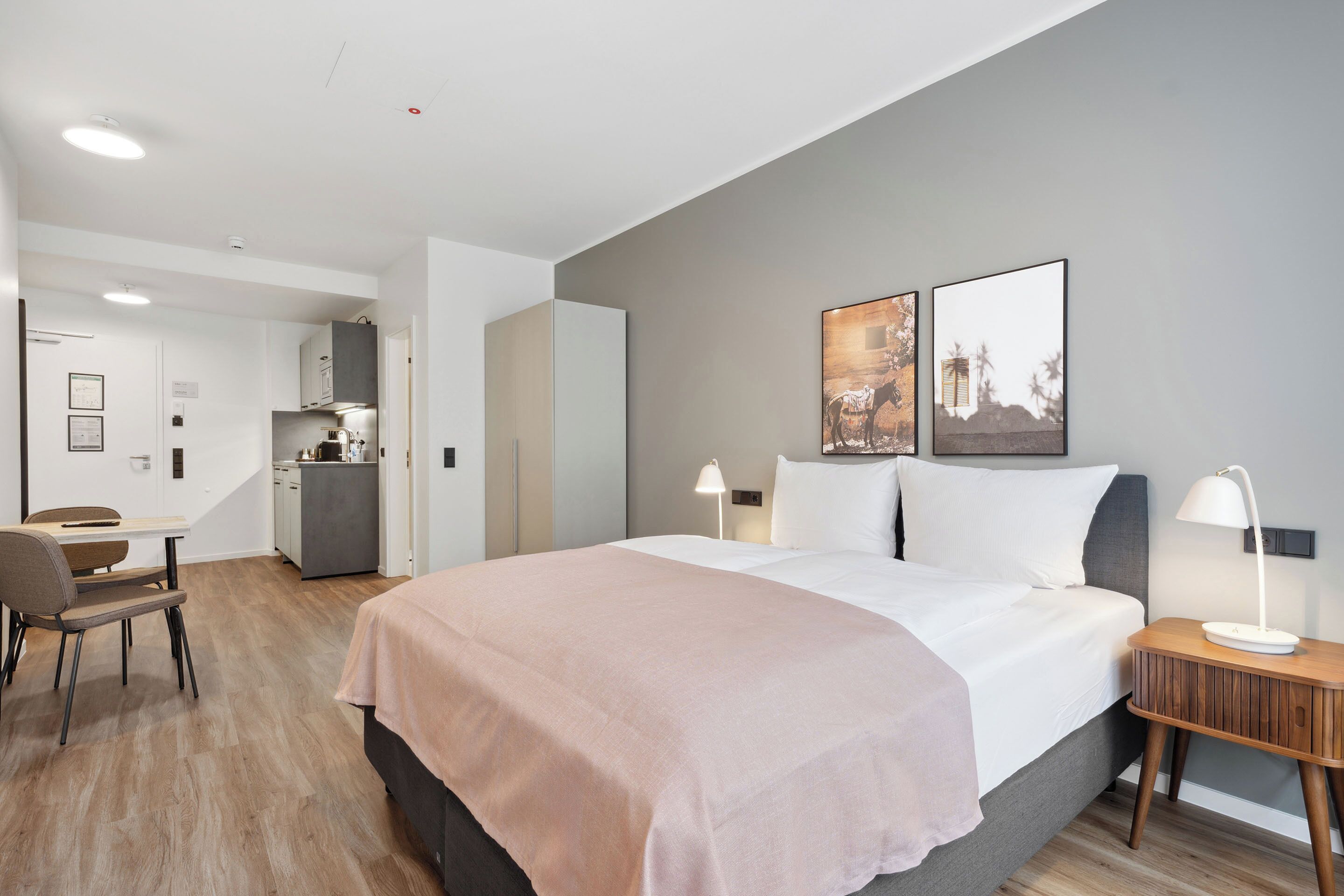 A modern hotel room with a large bed, bedside tables, a desk, and a kitchenette.