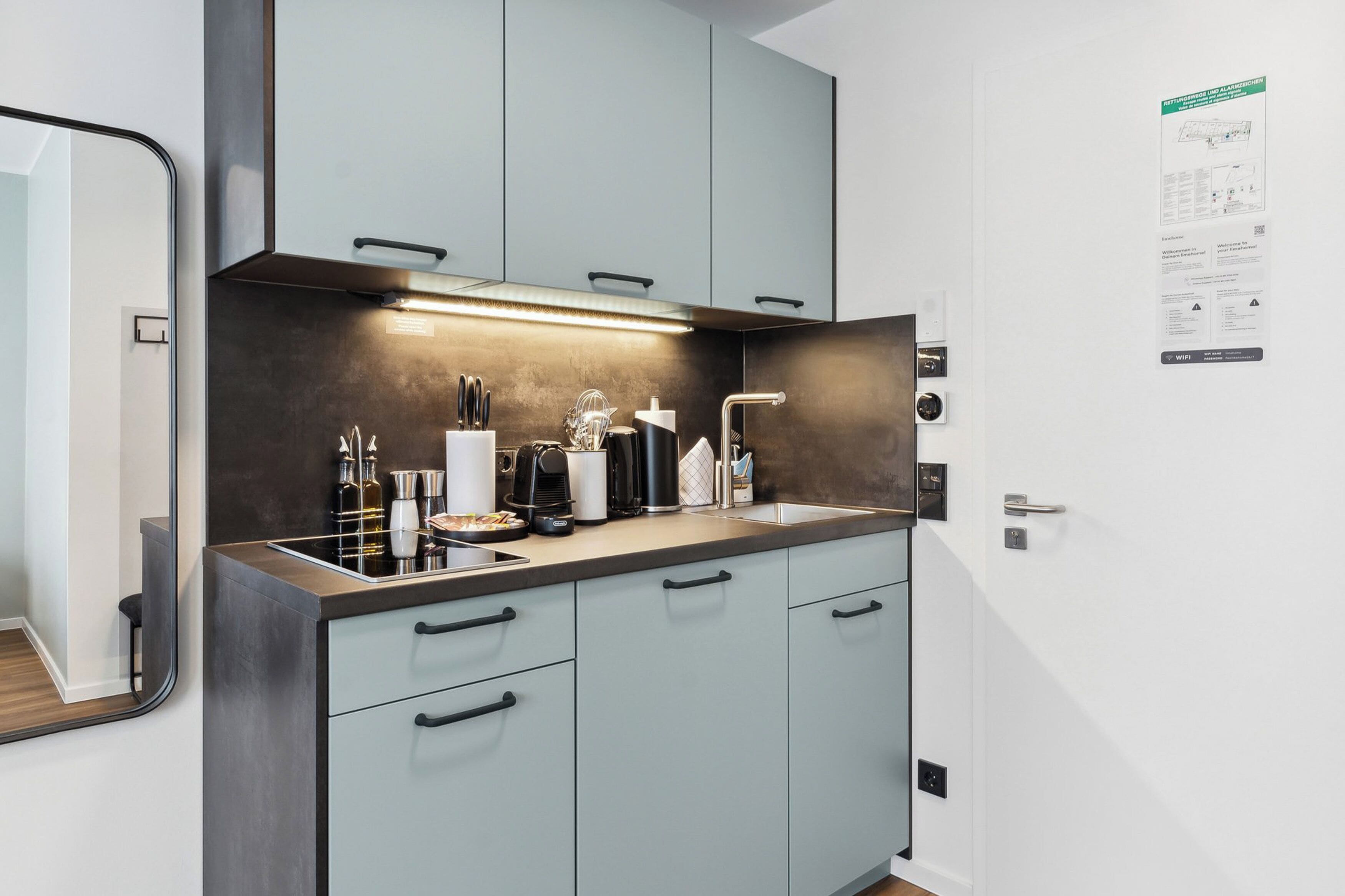 Eine moderne Küche mit hellblauen Schränken, einer Edelstahl-Arbeitsplatte und einem eingebauten Backofen. Ein Spülbecken mit Armatur ist vorhanden, und verschiedene Küchenutensilien und Geräte sind sichtbar.