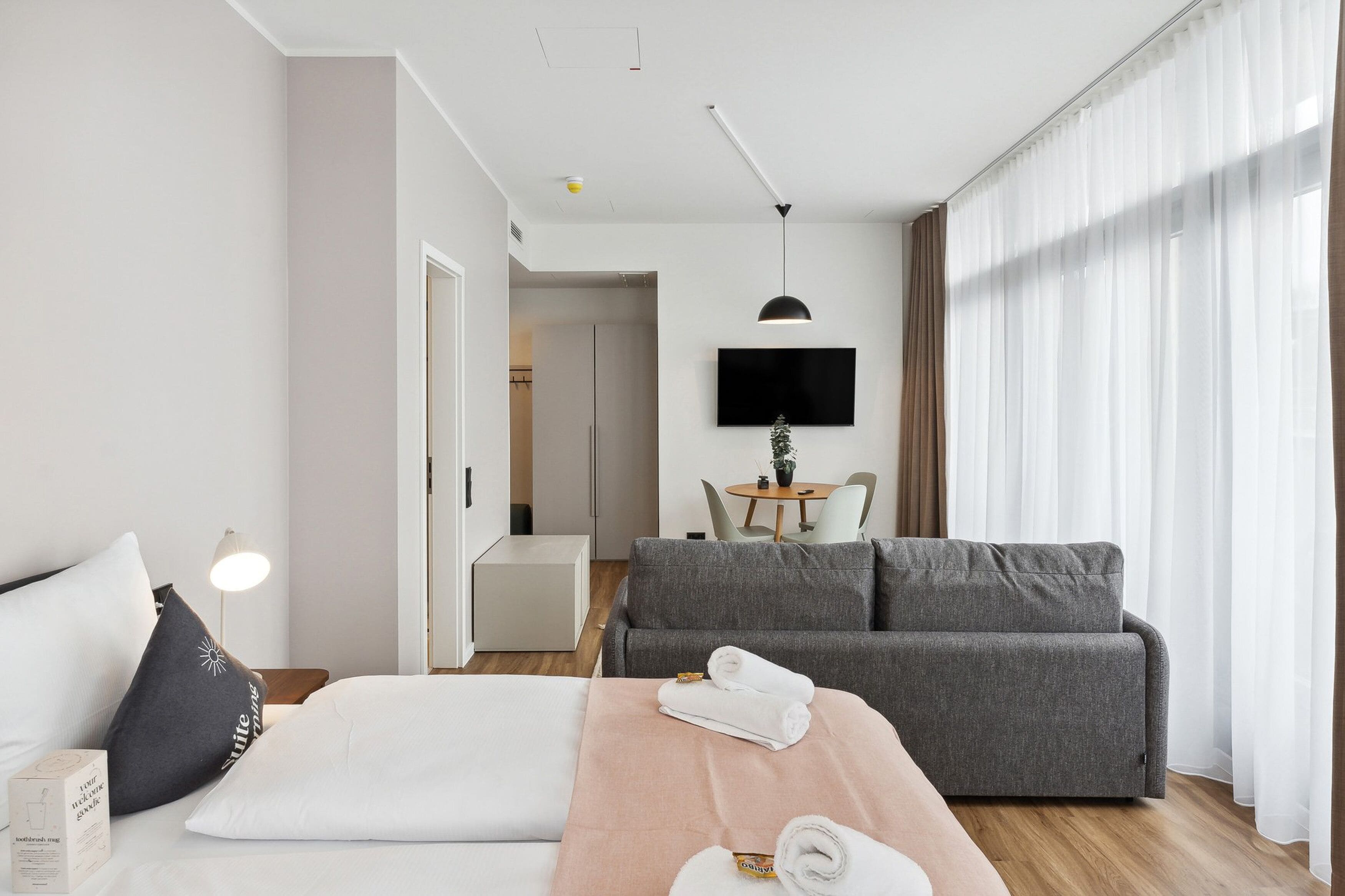 A modern hotel room with a bed, a grey sofa, a TV, and a dining area in the background.
