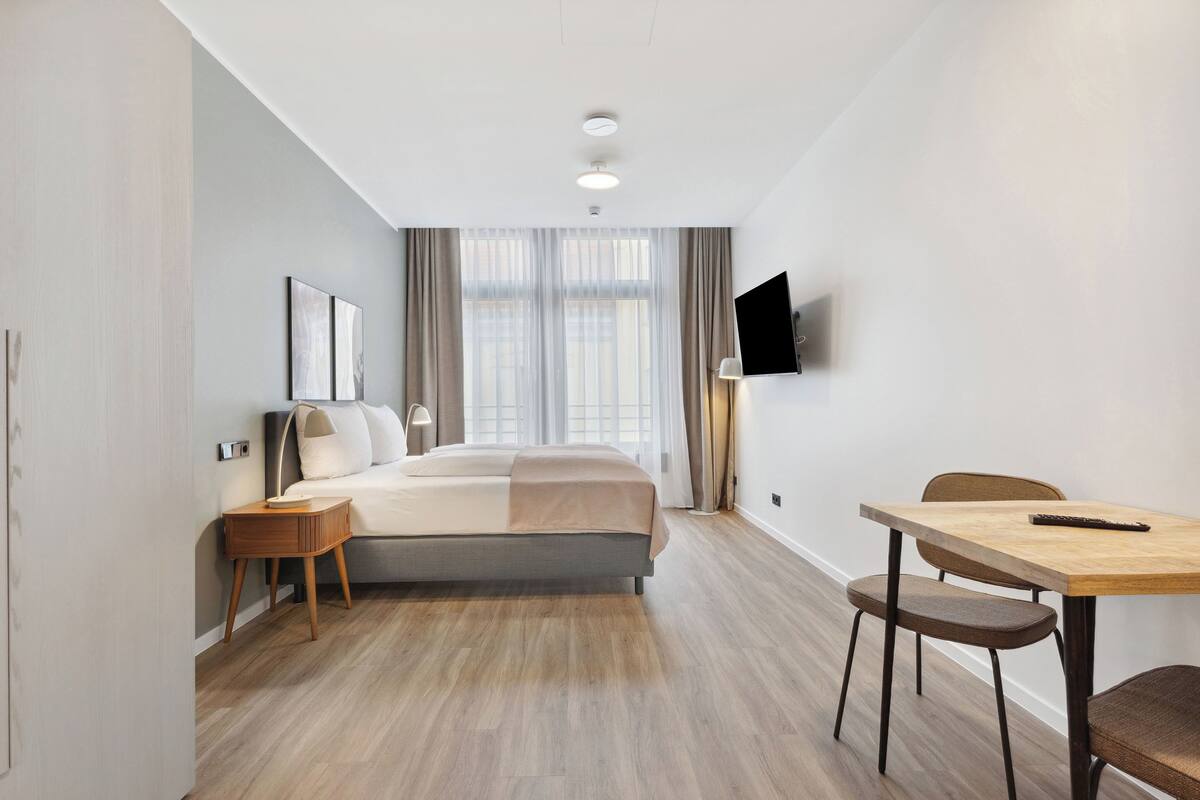 A modern hotel room with a bed, bedside table, lamp, TV, and a dining area with chairs and a table.