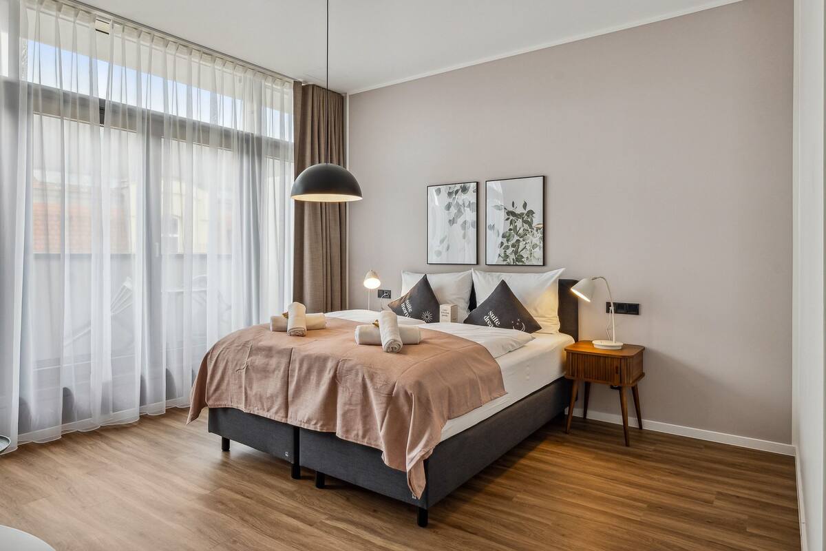 A modern bedroom with a large bed, sheer curtains, a hanging light, and two framed pictures on the wall.
