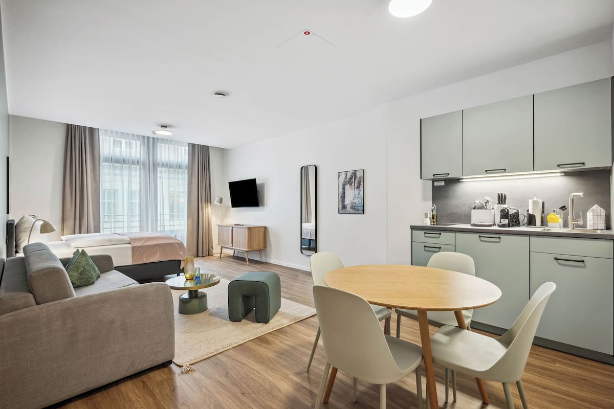 A modern hotel room with a bed, sofa, dining table, chairs, kitchenette, and a TV mounted on the wall.