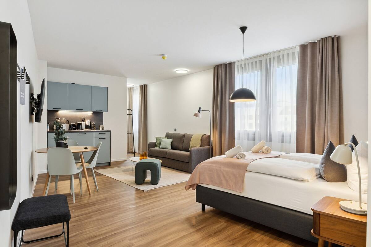 A modern hotel room with a bed, bedside table, lamp, and a kitchenette area.
