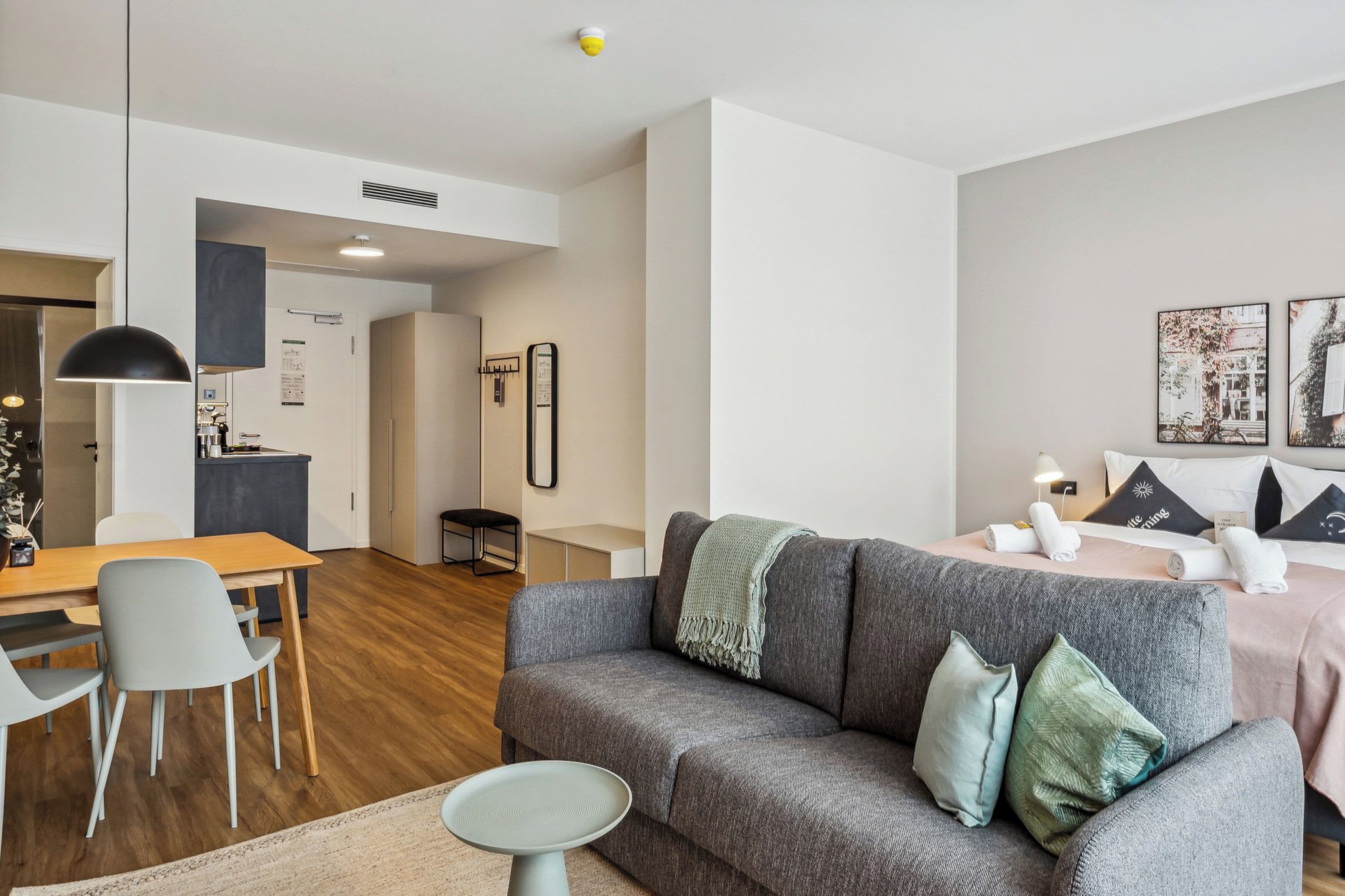 Ein modernes Wohnzimmer mit grauem Sofa, einem Esstisch mit Stühlen, einem kleinen runden Tisch und einer Küchenzeile im Hintergrund.