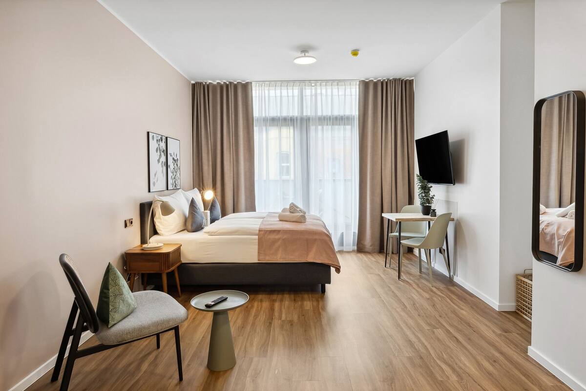 A modern hotel room with a large bed, a desk, a chair, a TV, and a window with curtains.