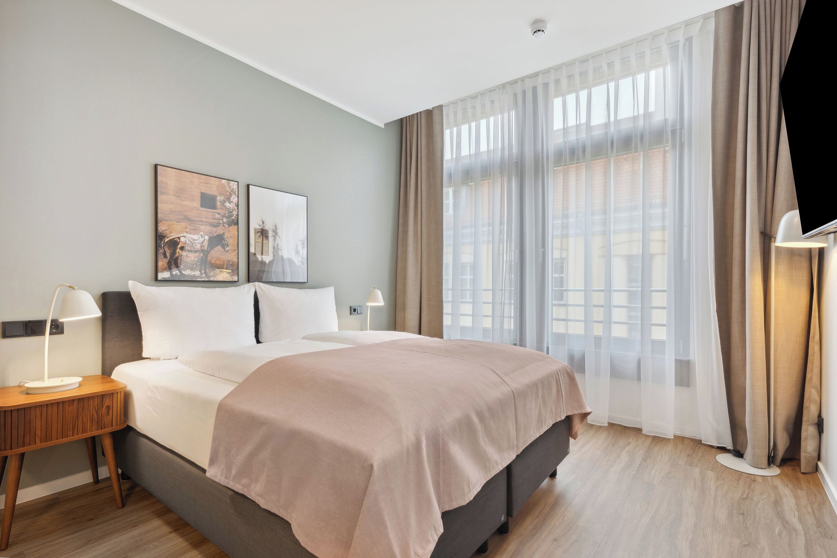 Una habitación de hotel moderna con una cama grande, una mesita de noche con lámpara y una ventana con cortinas translúcidas.