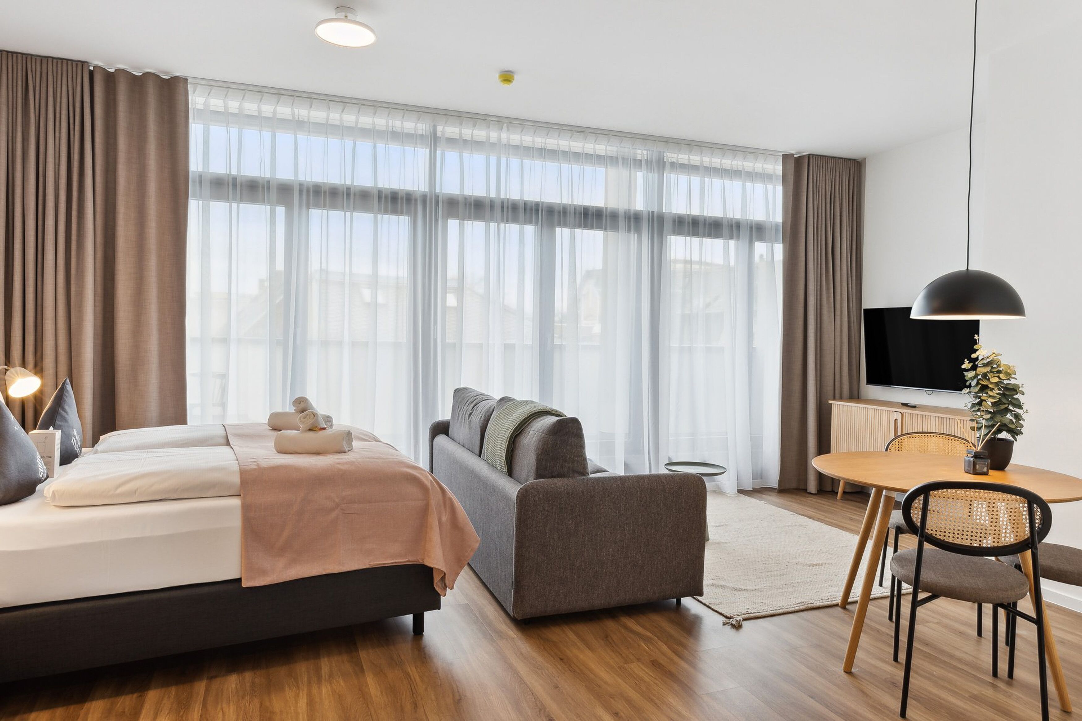 A modern hotel room with a bed, sofa, dining table, and TV.