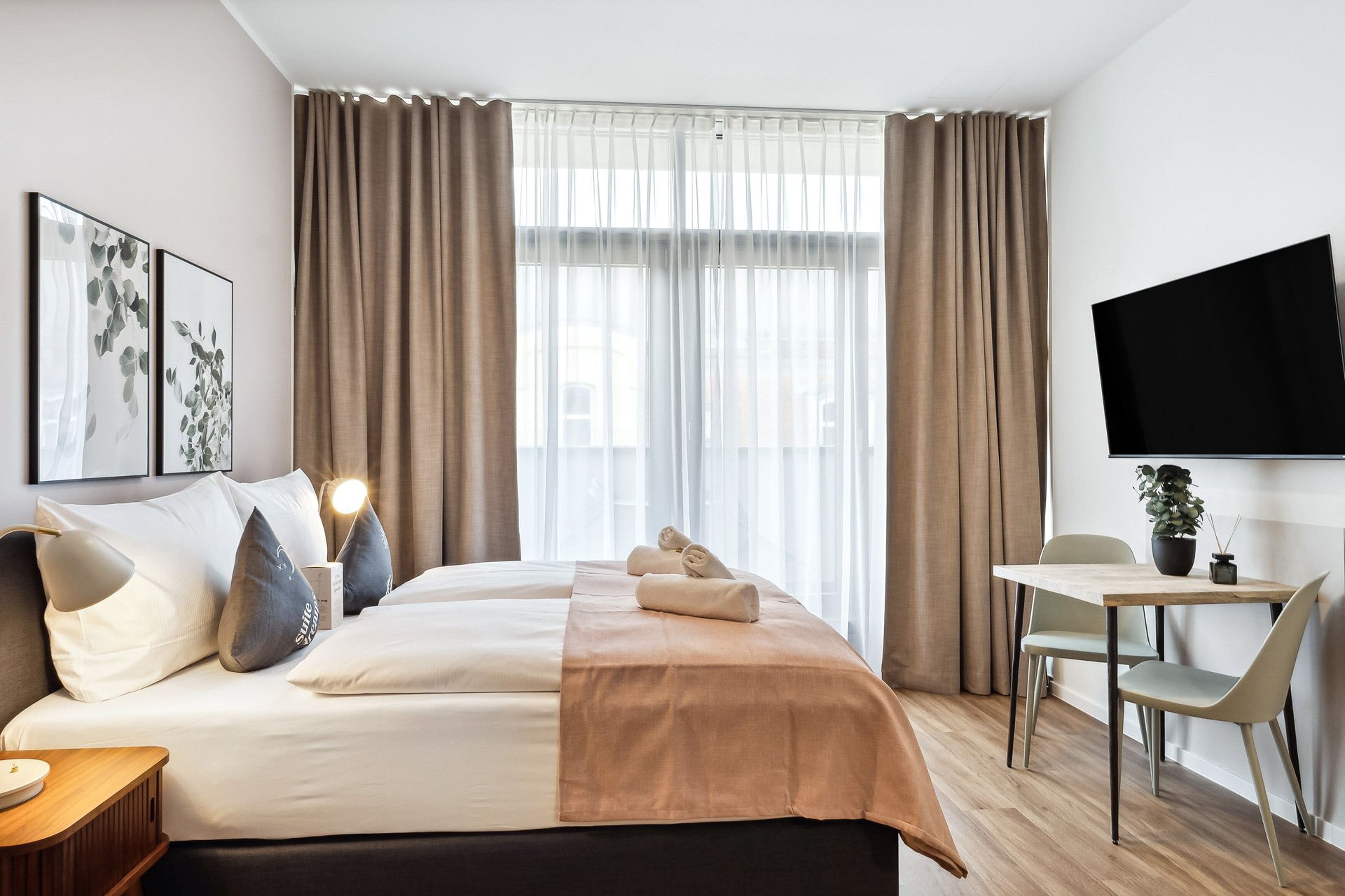 Ein Hotelzimmer mit Bett, Schreibtisch, Stuhl, Fernseher und einem Fenster mit Vorhängen.