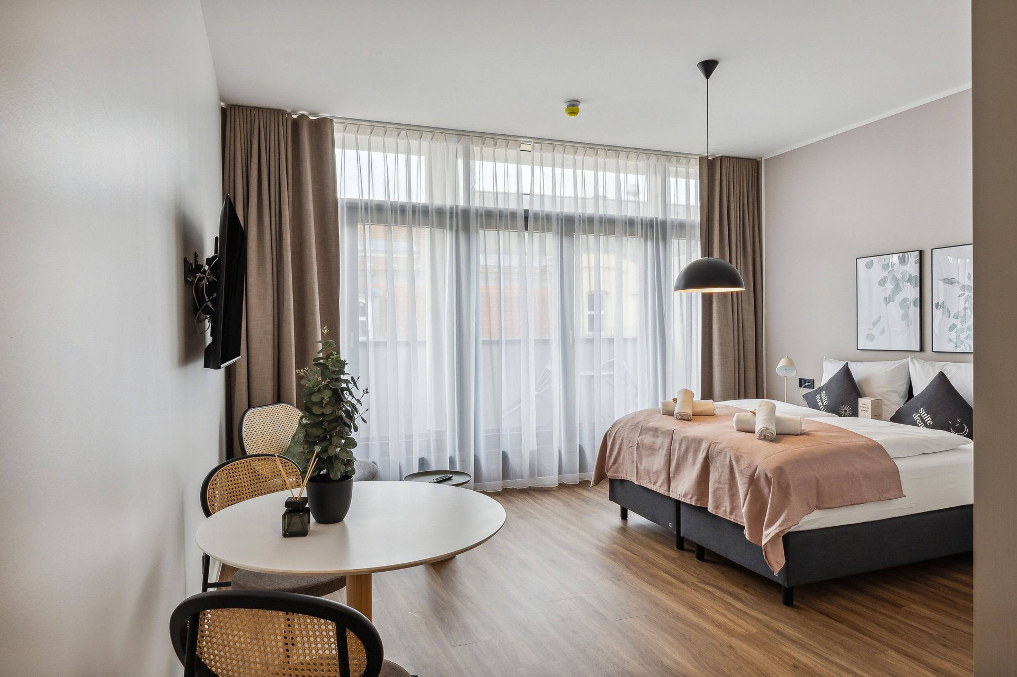 A modern hotel room with a bed, a dining table with chairs, a potted plant, and a large window with sheer curtains.