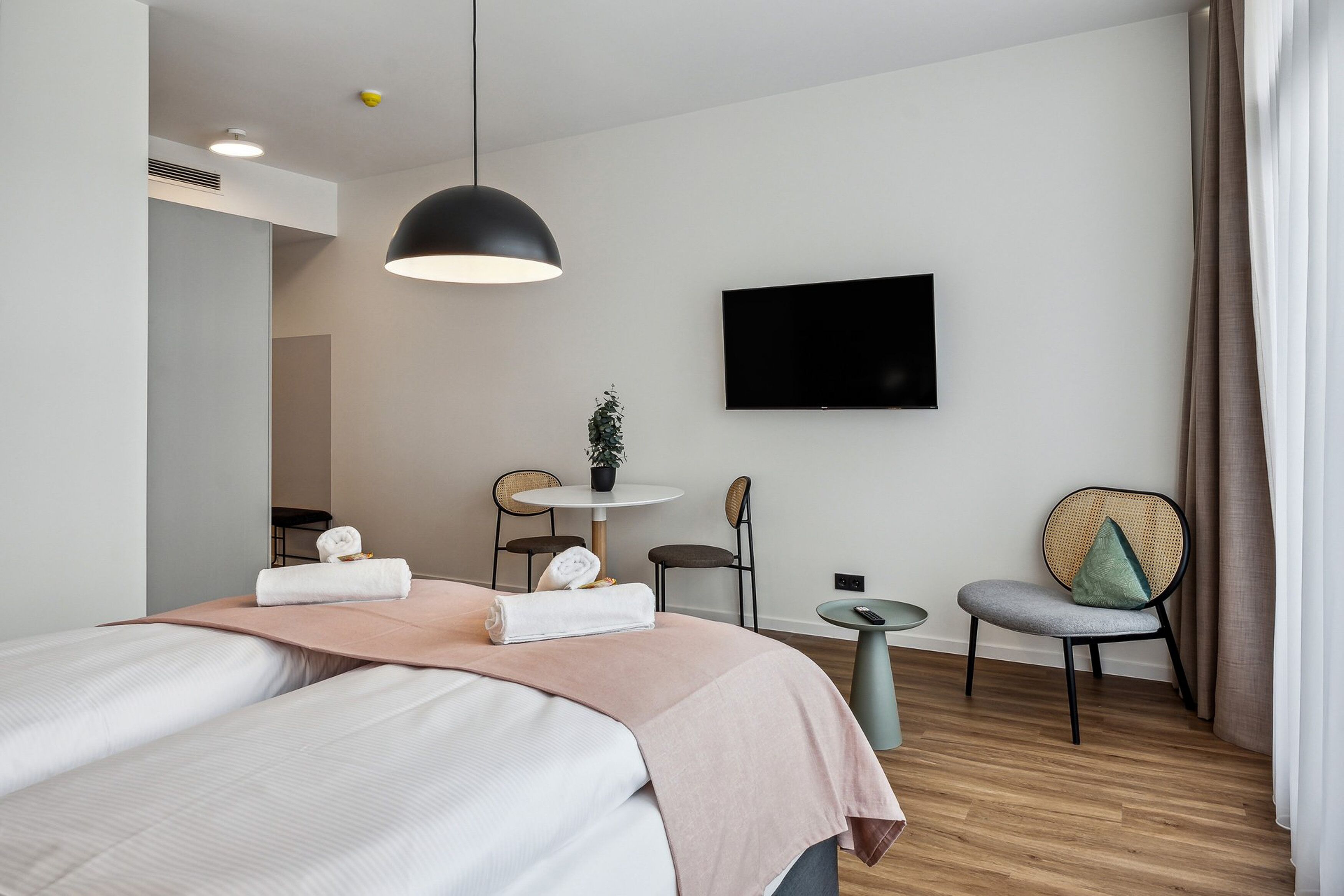 A modern hotel room with a bed, a round table, a chair, a television, and a small plant.