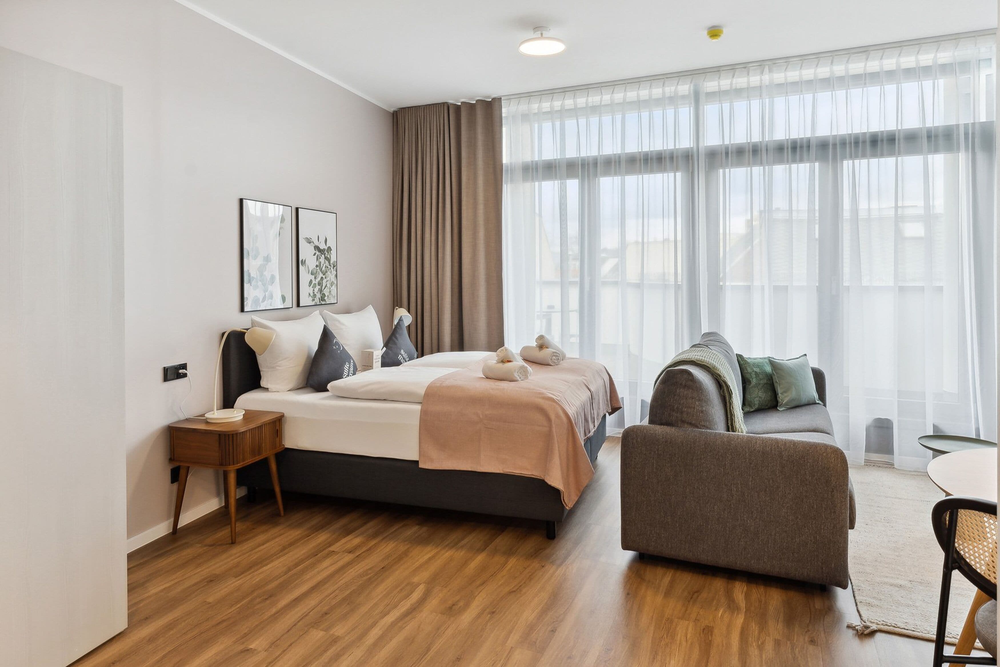 Quarto de hotel moderno com uma cama grande, um sofá, uma mesa pequena e uma janela ampla com cortinas.