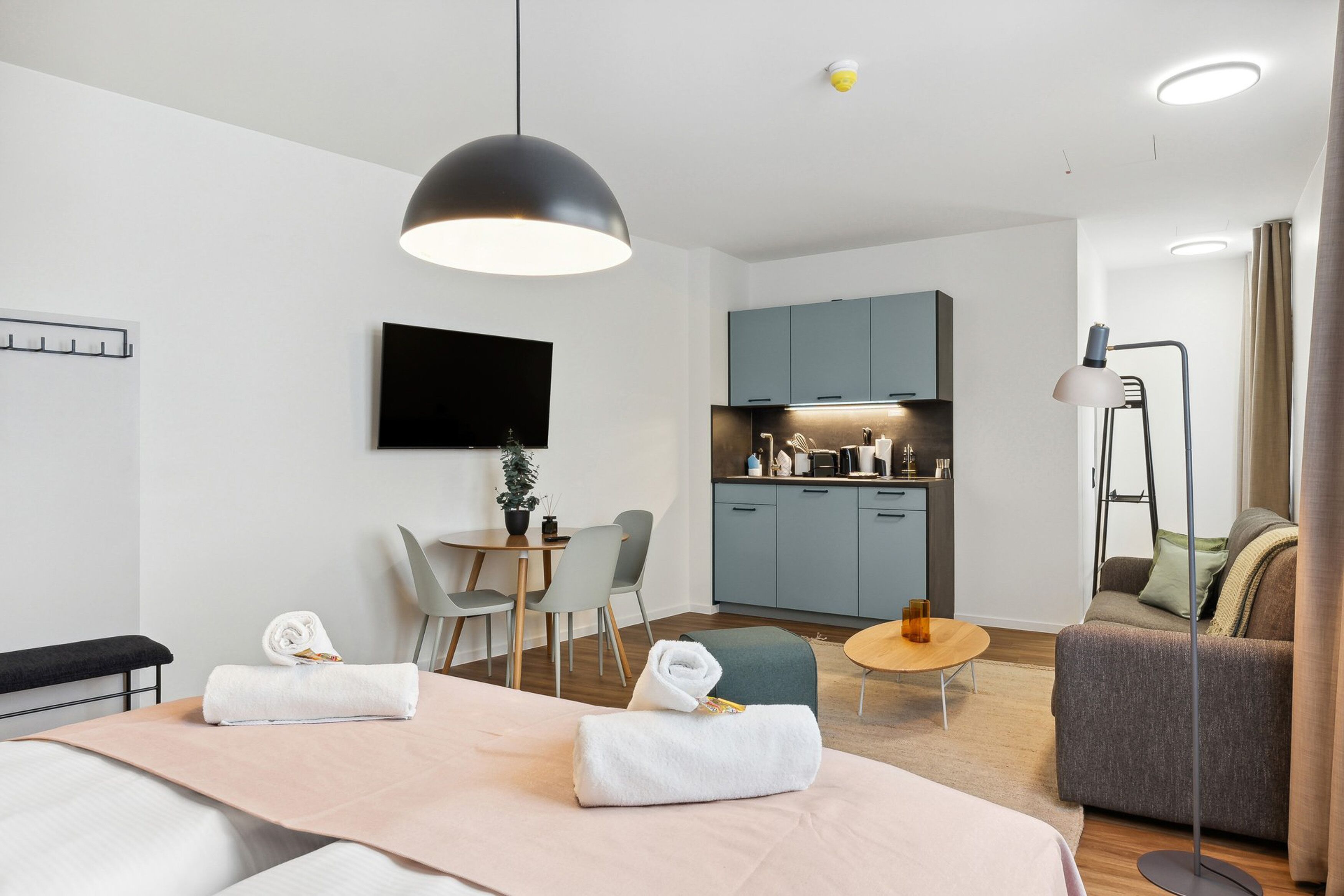 Una habitación de hotel moderna con cama, escritorio, silla, sofá, una mesita redonda pequeña, lámpara de pie y kitchenette.