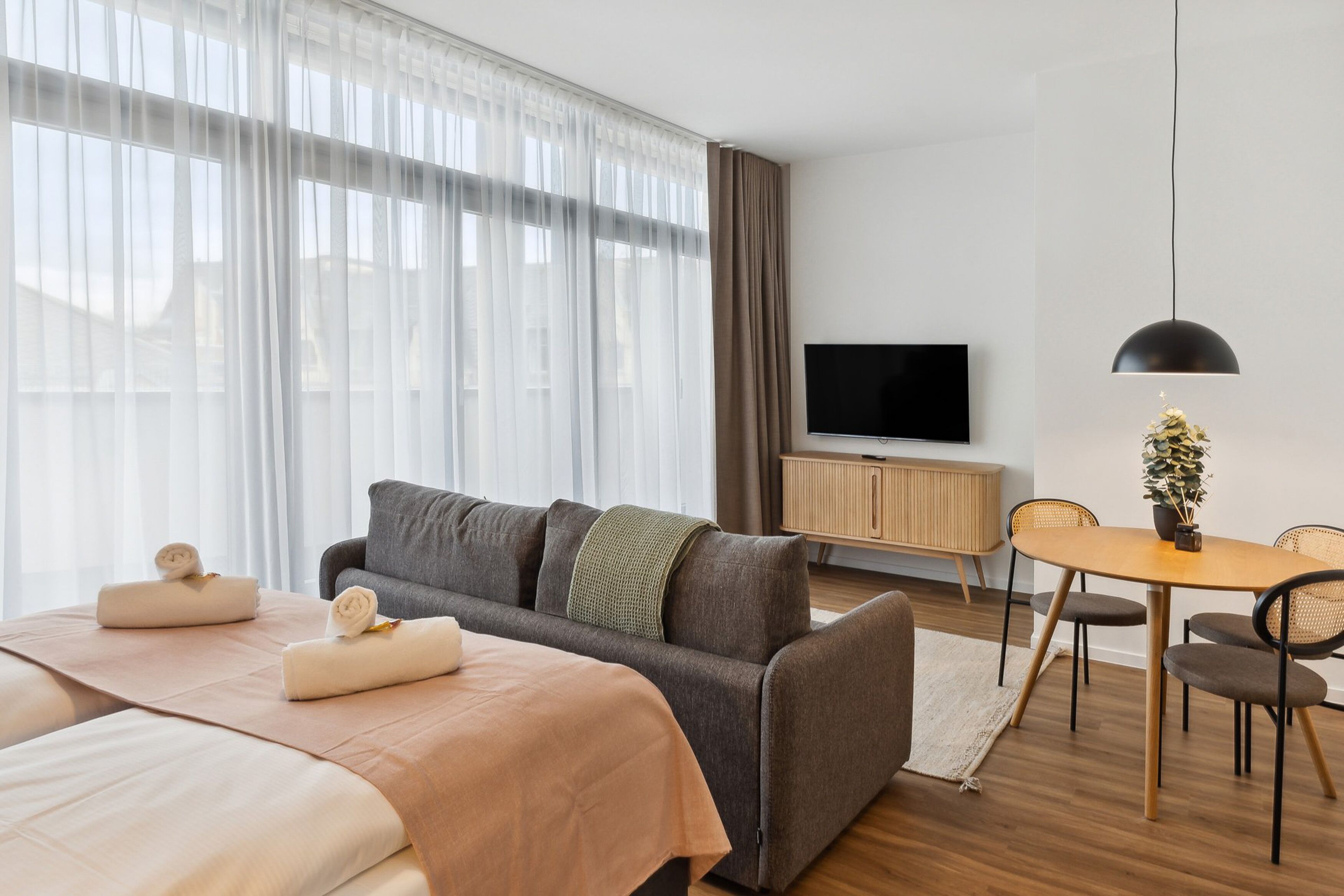 Habitació d'hotel moderna amb un llit gran, un sofà, una taula de menjador amb caires, una televisió i una planta en test.