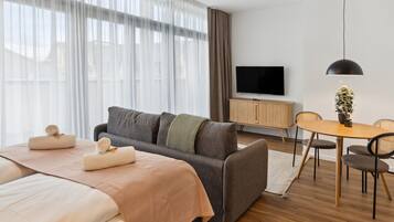Habitació d'hotel moderna amb un llit gran, un sofà, una taula de menjador amb caires, una televisió i una planta en test.