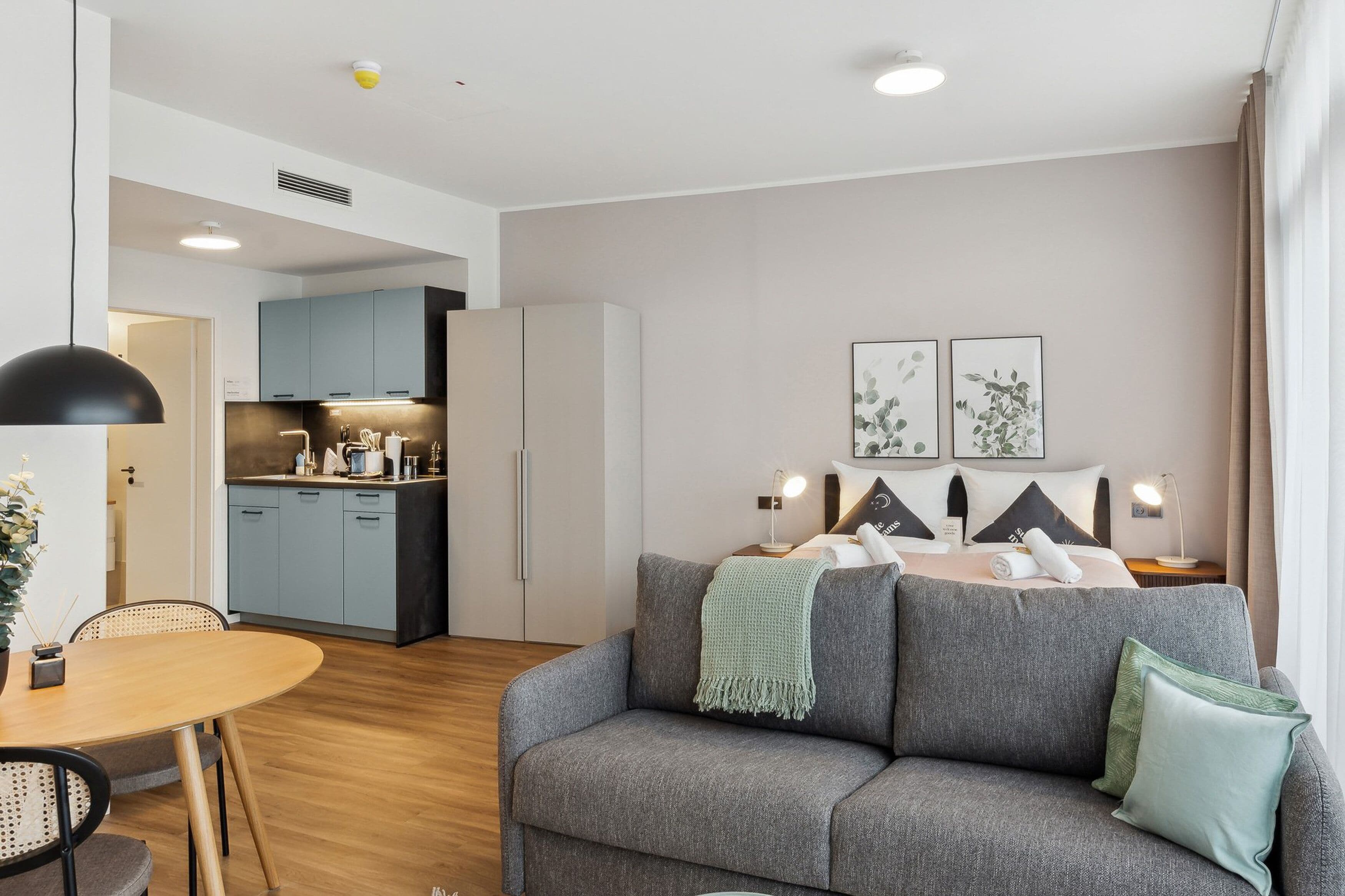 Una habitación de hotel moderna con un sofá gris, una mesa de comedor y una kitchenette al fondo.