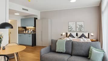 A modern hotel room with a grey sofa, a dining table, and a kitchenette in the background.