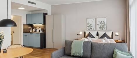 Una habitación de hotel moderna con un sofá gris, una mesa de comedor y una kitchenette al fondo.