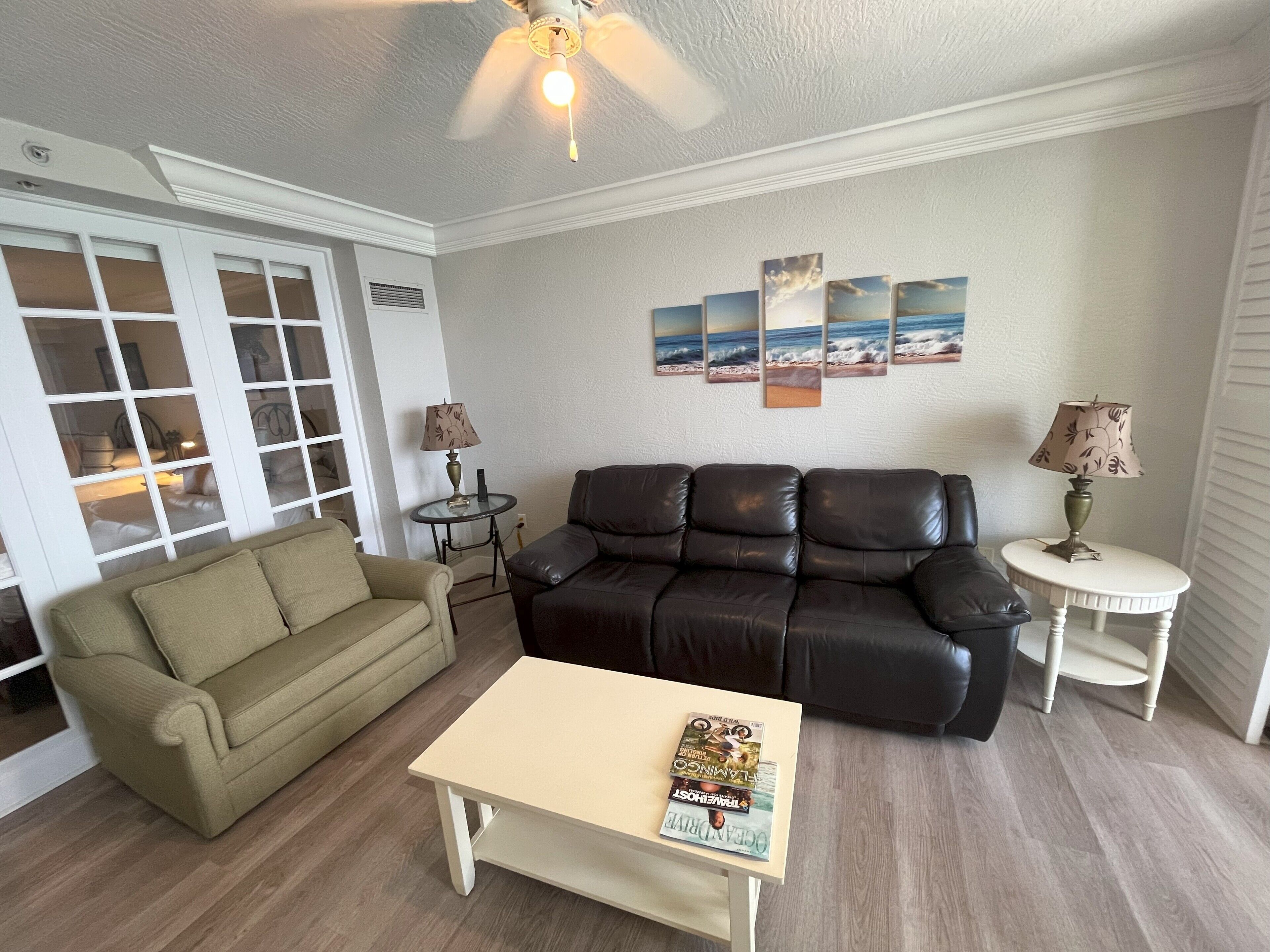 A living room with a beige sofa, a black leather couch, a coffee table with magazines, a small side table with a lamp, and a ceiling fan.
