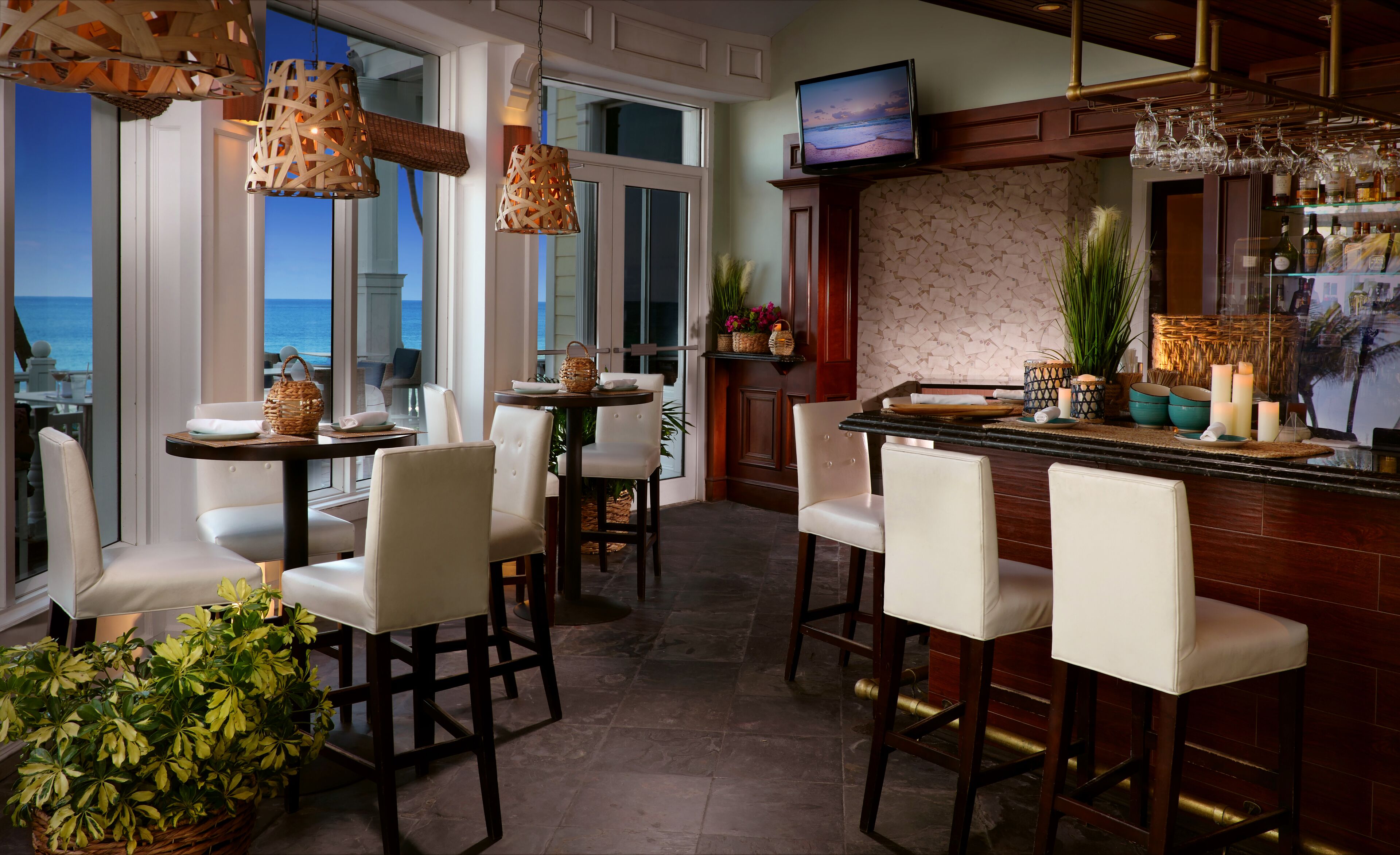 A bar area with a view of the ocean, wooden chairs, and a television screen.